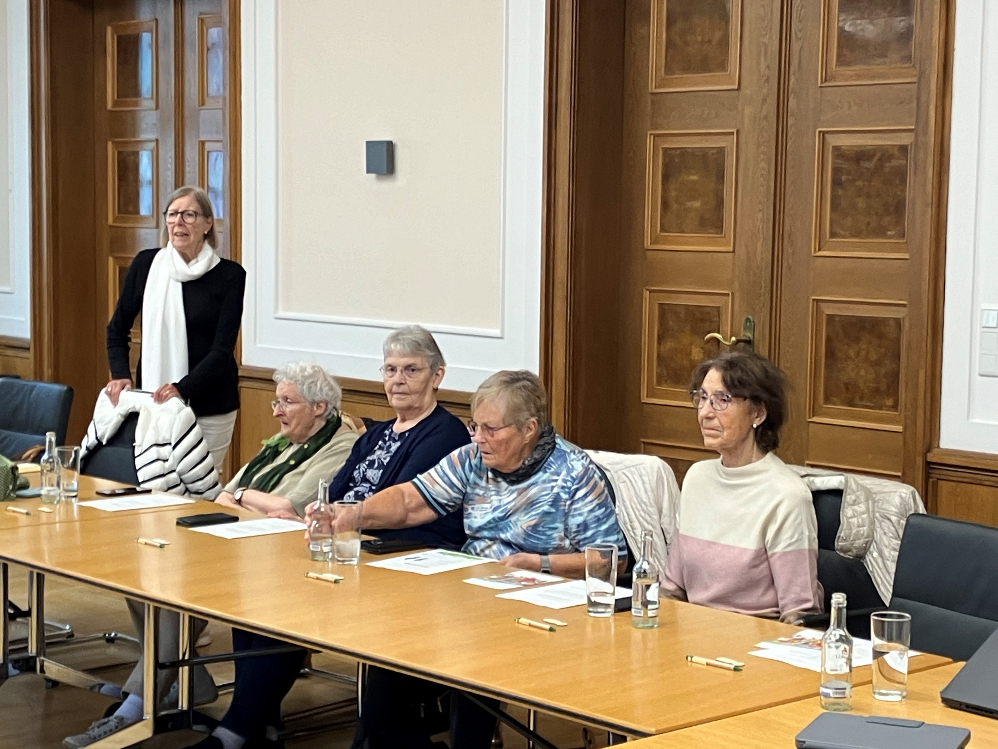 Vier Damen sitzen im großen Sitzungssaal an langen Holztischen nebeneinander, eine Dame steht und spricht zu den Gästen. Im Hintergrund sind die schweren Holztüren des Rathaussaales zu erkennen.