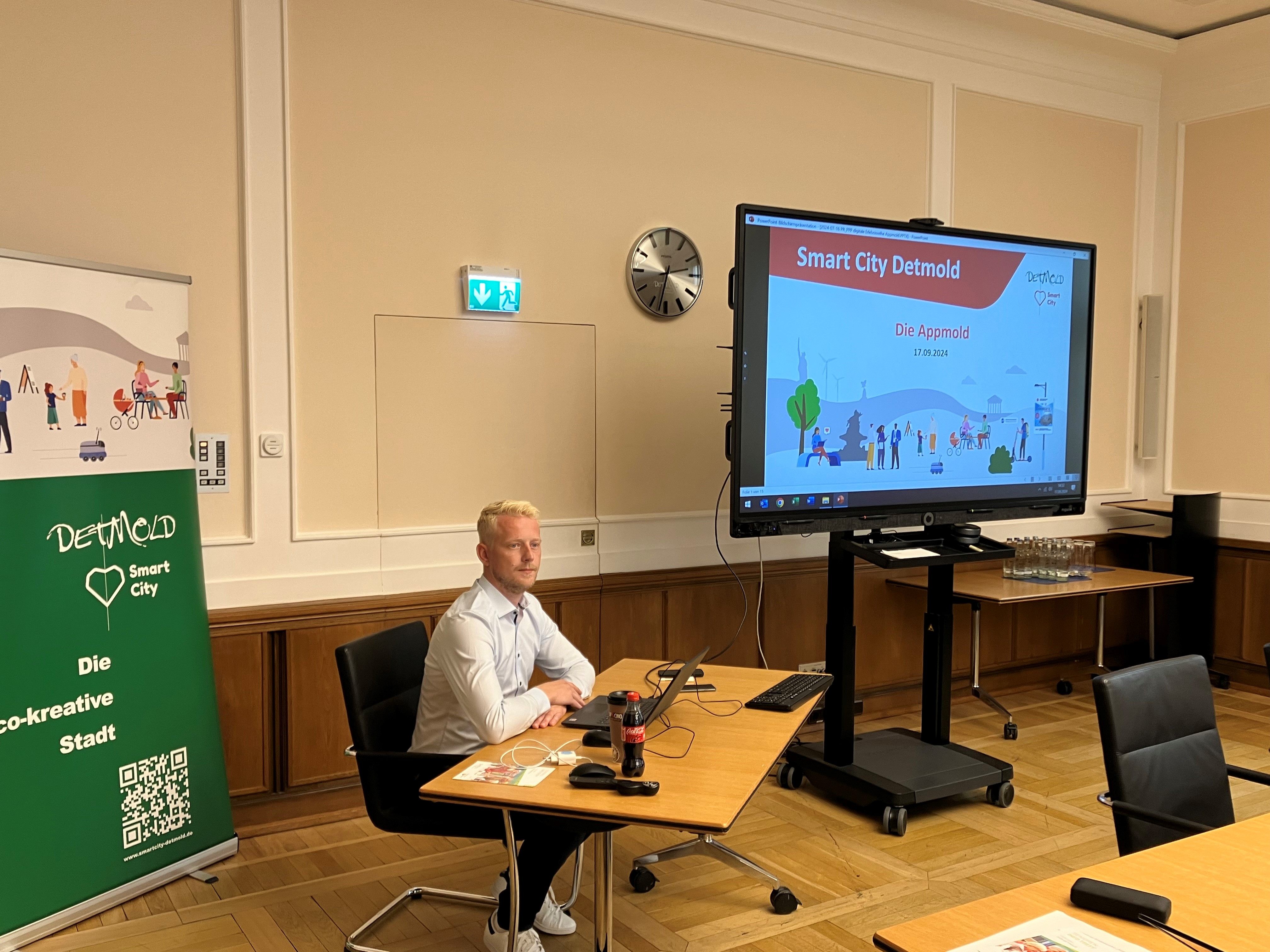 Ein blonder Mitarbeiter der Stadt sitzt hinter einem Schreibtisch im Großen Sitzungssaal des Rathauses in Detmold. Im Hintergrund ist links ein Banner der Smart City Detmold zu sehen, rechts im Bild ein großer Bildschirm auf Rollen mit der Startseite der Präsentation "Appmold". 
