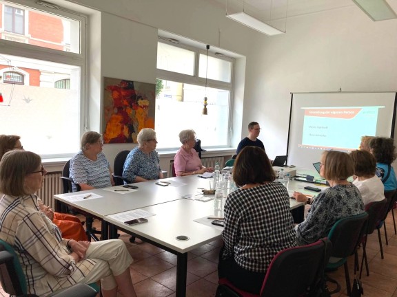 In einem großen Raum sieht man Seniorinnen und Senioren an einem Tisch itzen. Oben links sitzt ein junger Mann, der die Menschen über die Stadtapp informiert. 