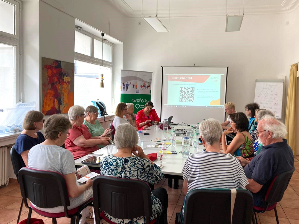 Das Foto zeigt eine Aufnahme aus dem Hintergrund des Raumes, in dem die Seniorenschulung stattfindet. 15 Personen sitzen an einem großen weißen Tisch und bedienen ihre Mobiltelefone.