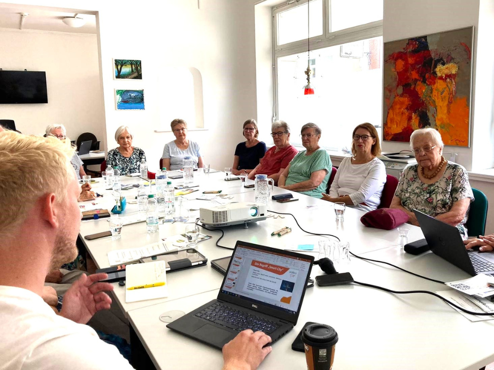 Das Foto zeigt einen voll bestzten Raum mit 14 Senior*innen, die an einem großen weißen Tisch sitzen und dem  Referenten (angeschnitten von hinten, vorne im Bild zu sehen). 
