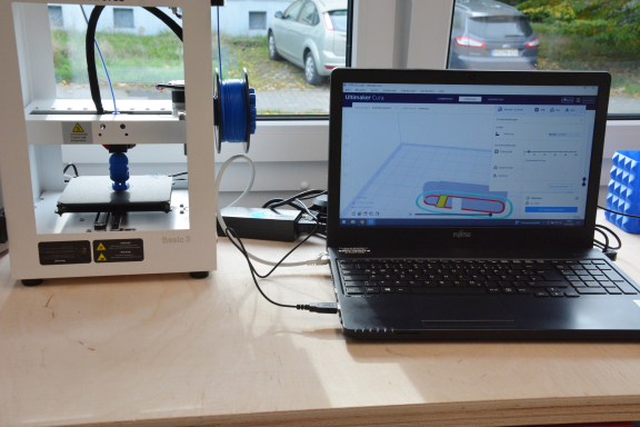 Das Foto zeigt rechts auf einem Tisch stehend ein aufgeklapptes Laptop. Links daneben steht ein weißer 3D-Drucker. 