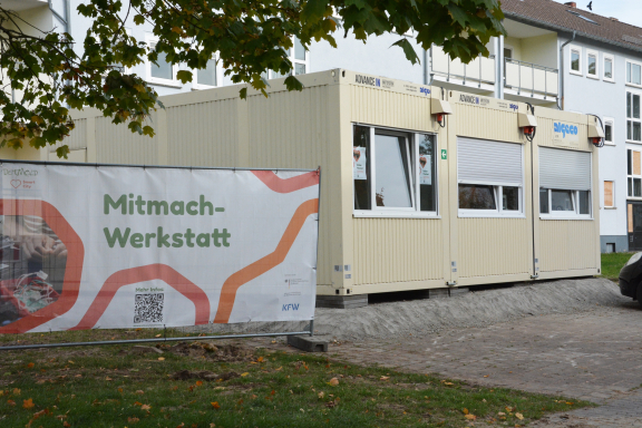 Außenaufnahme der Mitmachwerkstatt: Man sieht drei verbundene beigefarbene Container mit Fenstern, links daneben hängt ein Plakat an einem Bauzaun 