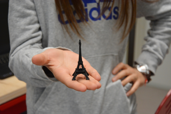 Ein junges Mädchen balanciert einen vom 3D-Drucker ausgedruckten Eifelturm auf ihrer Hand und streckt diesen Richtung Kamera. Sie trägt einen grauen Pullover. Ihr Gesicht  ist nicht zu sehen. 