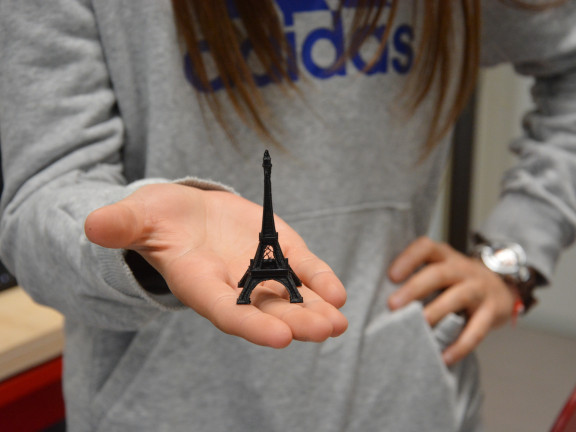 Ein junges Mädchen balanciert einen vom 3D-Drucker ausgedruckten Eifelturm auf ihrer Hand und streckt diesen Richtung Kamera. Sie trägt einen grauen Pullover. Ihr Gesicht  ist nicht zu sehen. 