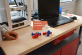 Auf einer Werkbank mit heller Holzplatte stehen vier ausgedruckte 3D-Modelle. Man erkennt ein rotes Boot, zwei Fischskeleltte (je eines in Blau und Rot) sowie einen blauen, kleinen Astronauten. 