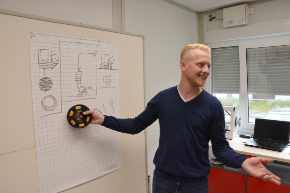 Ein junger Mann im blauen Pullover präsentiert eine Filamentrolle. Er steht vor einem Flipchart. 