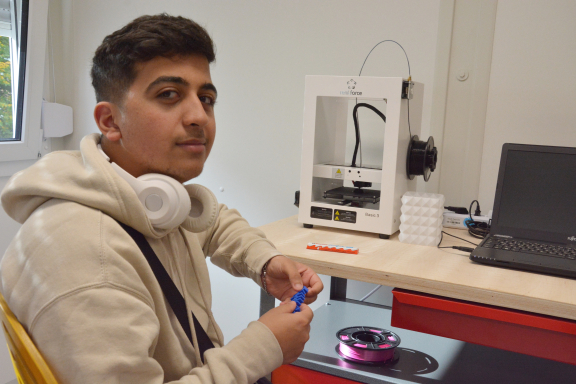 Ein junger Mann mit Schnäuzer trägt einen weißen Hoodie und sitzt an einer Werkbank und blickt in die Kamera. In der Hand hält er einen Audruck aus dem 3D-Drucker. 