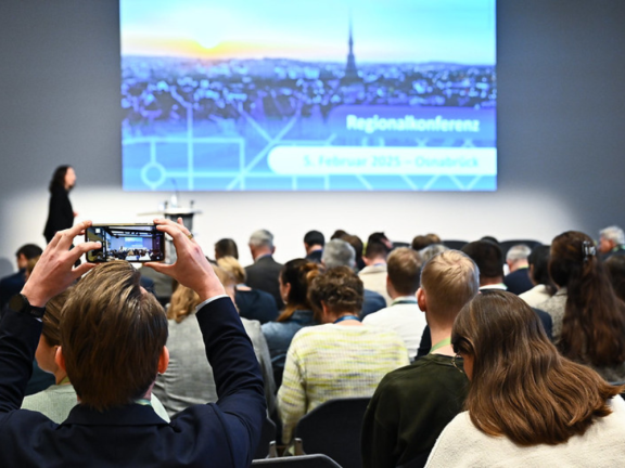 Das Foto wurde von der Rückseite des Raumes aufgenommen und zeigt eine Momentaufnahme eines Bühnenvortrages. Im Bild sind die Köpfe des Publikums in mehreren Reihen im Vordergrund zu sehen. 