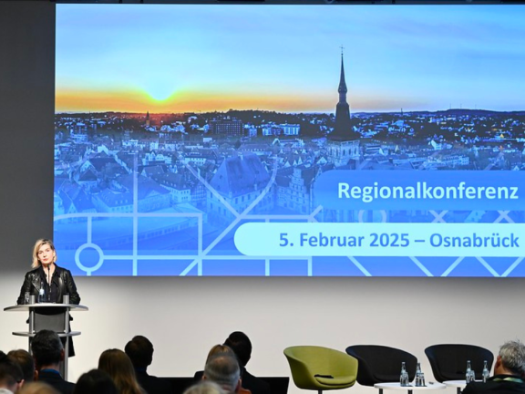 Das Bild zeigt Renate Mitterhuber auf der Bühne beim MPSC-Regionalkongress in Osnabrück. Sie steht hinter einem Pult, im Hintergrund ist eine blaue Folie zu sehen auf der 