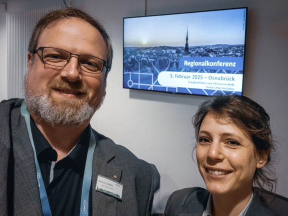 Ein Mann mit Bart und Brille und eine junge Frau sind großformatig auf dem Foto zu sehen und lächeln in die Kamera. Im Hintergrund sieht man einen Bildschirm, der auf die Regionalkonferenz am 5. Februar 2025 in Osnabrück hinweist. 