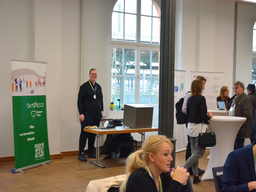 Das Foto zeigt den Ausstellungsraum, vorne sitzt (angeschnitten) eine Frau an einem Tisch. Im Hintergund steht ein junger Mann an einem Tisch mit dem QR-Code-Würfel. Neben ihm sieht man einen Banner.