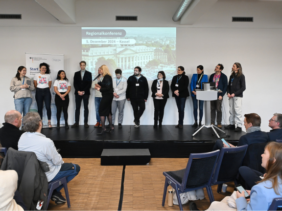 Man sieht eine große Bühne auf der 12 Personen stehen und nacheinander präsentieren. 