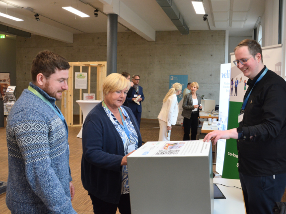 Ein Mann und eine Frau stehen vor einem Tisch mit einem QR-Code-Würfel und sind mit dem jungen Mann, Mitarbeiter der Stadt Detmold, im Gespräch. Alle lächeln. 