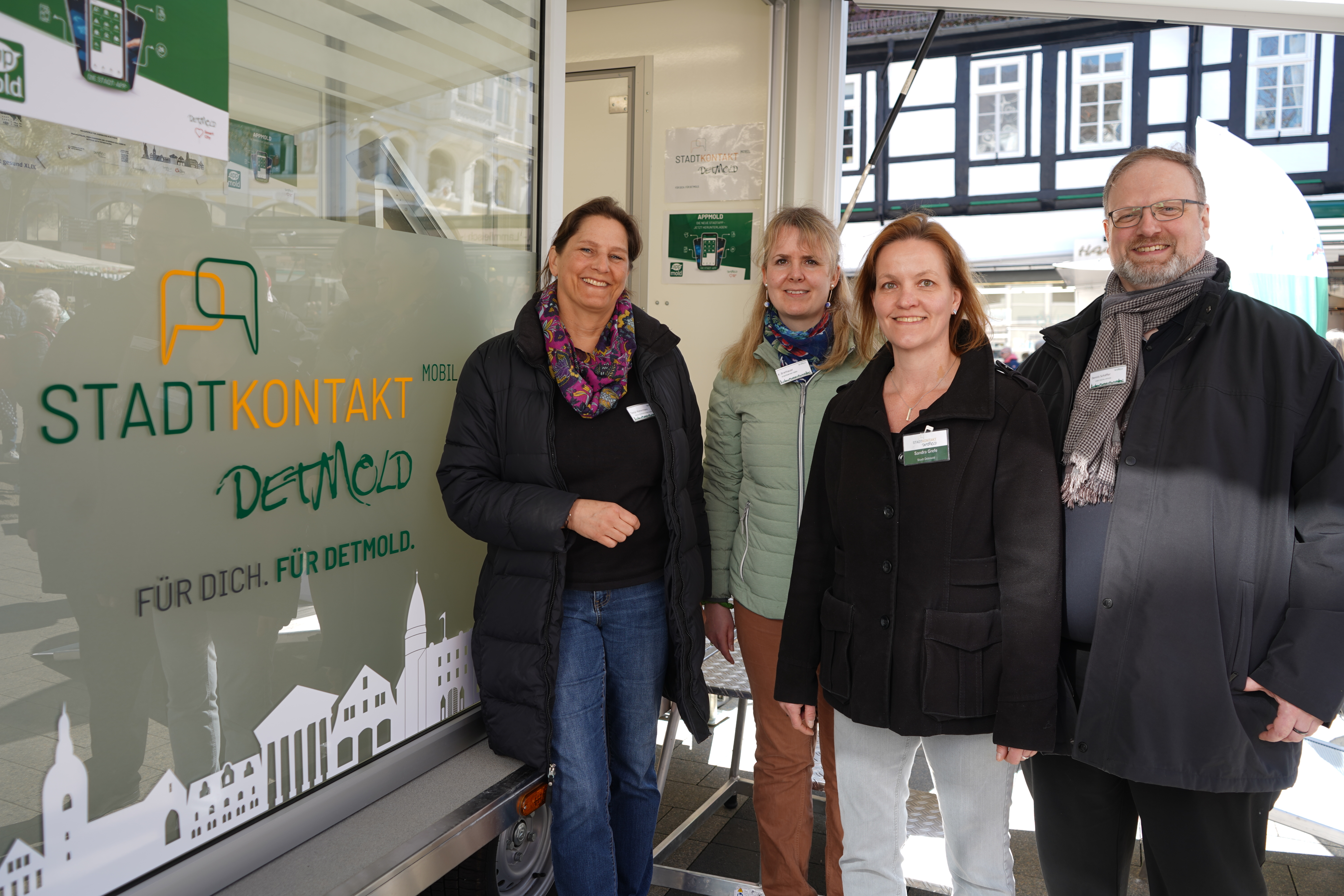 Das Bild zeigt vier Mitarbeiter*innen der Stadt Detmold, vor dem städtischen Anhänger namens "Stadtkontakt mobil. Vom Anhänger sind lediglich die große Glasfront sichtbar, vor der die Menschen stehen.