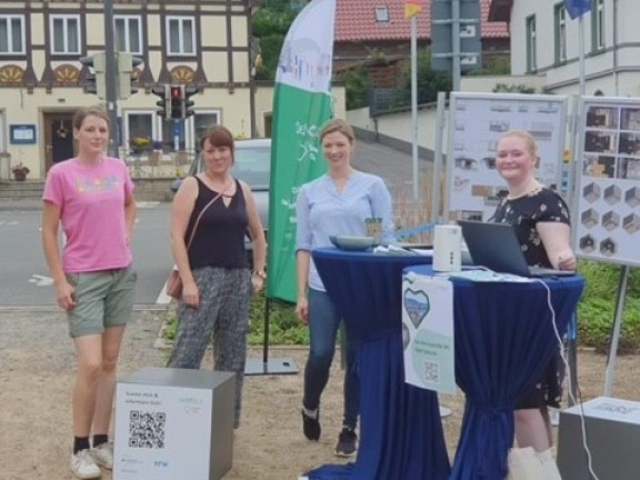 Das Foto zeigt vier junge Frauen, die im Halbkreis an zwei Stehtischenn mit blauen Hussen stehen. Im Hintegrund sieht man ein gelb-braunes Fachwerkhaus und eine grün-weiße Beachflag. 