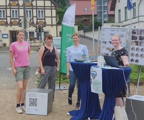 Das Foto zeigt vier junge Frauen, die im Halbkreis an zwei Stehtischenn mit blauen Hussen stehen. Im Hintegrund sieht man ein gelb-braunes Fachwerkhaus und eine grün-weiße Beachflag.