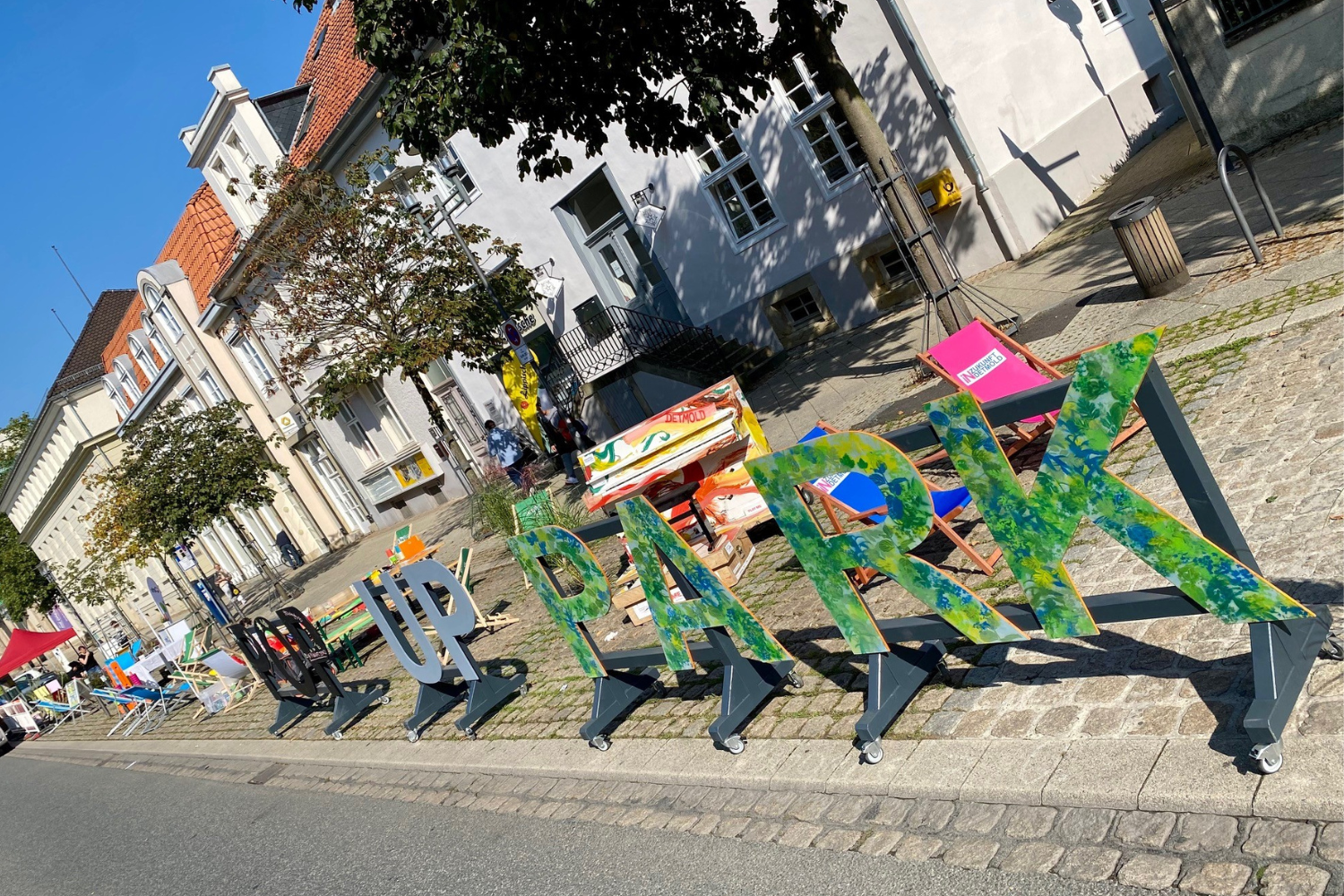 Das Foto zeigt eine Straße, bei der die Parkplätze in Aktionsflächen verwandelt wurden. Im Vordergund sieht man die bunt bemalten Großbuchstaben POP UP PARK