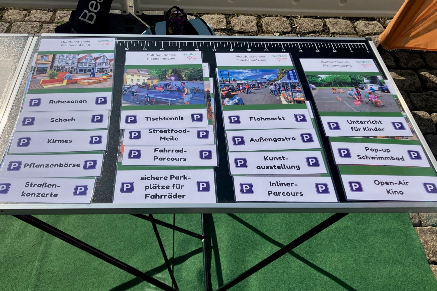 Man sieht einen schwarzen Tapeziertisch, fotografiert von oben, im Hintergrund sieht man Kopfsteinpflaster. Auf dem Tisch liegen über 10 DIN a4 Folien, die Alternativnutzungen für Parkplätze symbolisieren. Zu sehen sind unter anderem Begriffe (unterstützt durch Symbolfotos) wie Straßenkonzerte, Kirmes, Schach, Streetfoodmile, Tischtennis, Außengastro, Flohmarkt sowie zahlreiche weitere.