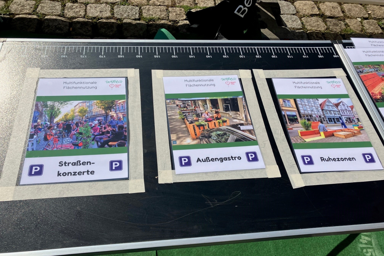 Man sieht einen schwarzen Tapeziertisch, fotografiert von oben, im Hintergrund sieht man Kopfsteinpflaster. Auf dem Tisch sieht man drei abgeklebte Flächen, die Parkplätze symbolisieren. Teilnehmer können mit Karten in DIN A4 Größe ihre Lieblingsnutzung für Parkplätze als Stimme abgeben. Zu sehen: Sttraßenkonzerte, Außengastronomie und Ruhezonen.