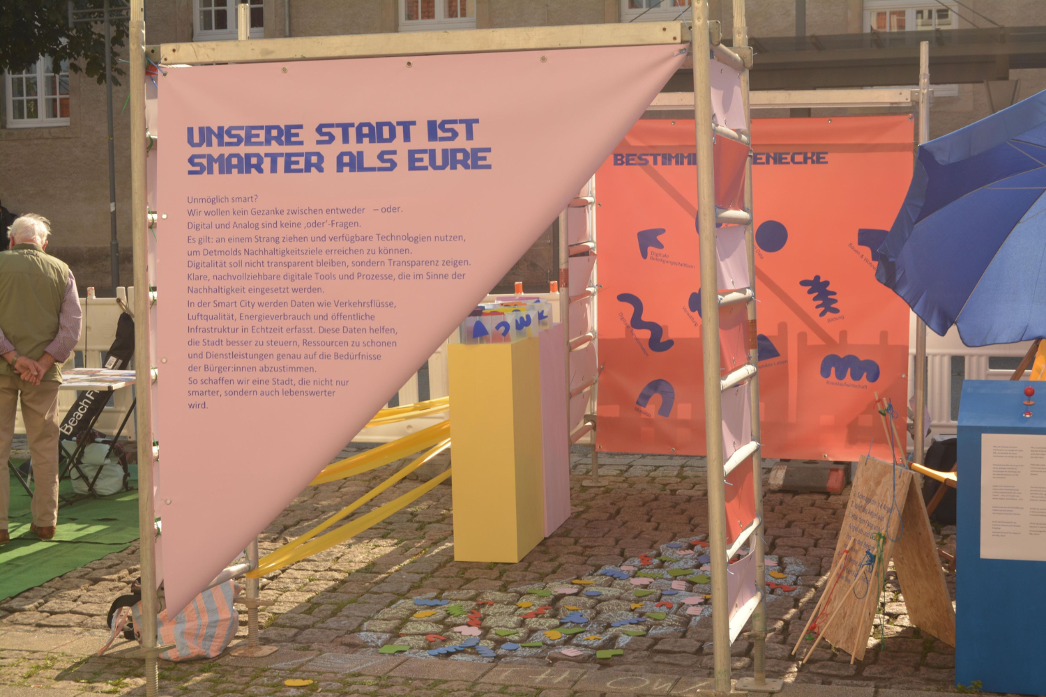 Das Foto zeigt eine Installation auf einer der Parkflächen. Ein etwa 2,5 Meter hohes Gerüst wurde auf der Parkfläche aufgebaut, bunte Plakate hängen an den Gerüsten. Die Plakate erklären die Bürgerbeteiligung, die hier erprobt werden soll. Man kann die Schrift (bis auf die Überschrift "Unsere Stadt ist smarter als eure") nicht erkennen.