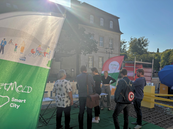 Vorne rechts im Bild sieht man eine grün-weiße Beachflag der Detmold Smart City ins Foto ragen. 5 Besucher*innen informieren sich an einem Tisch zum Projekt bei einer Mitarbeitenden der Stadt Detmold. 