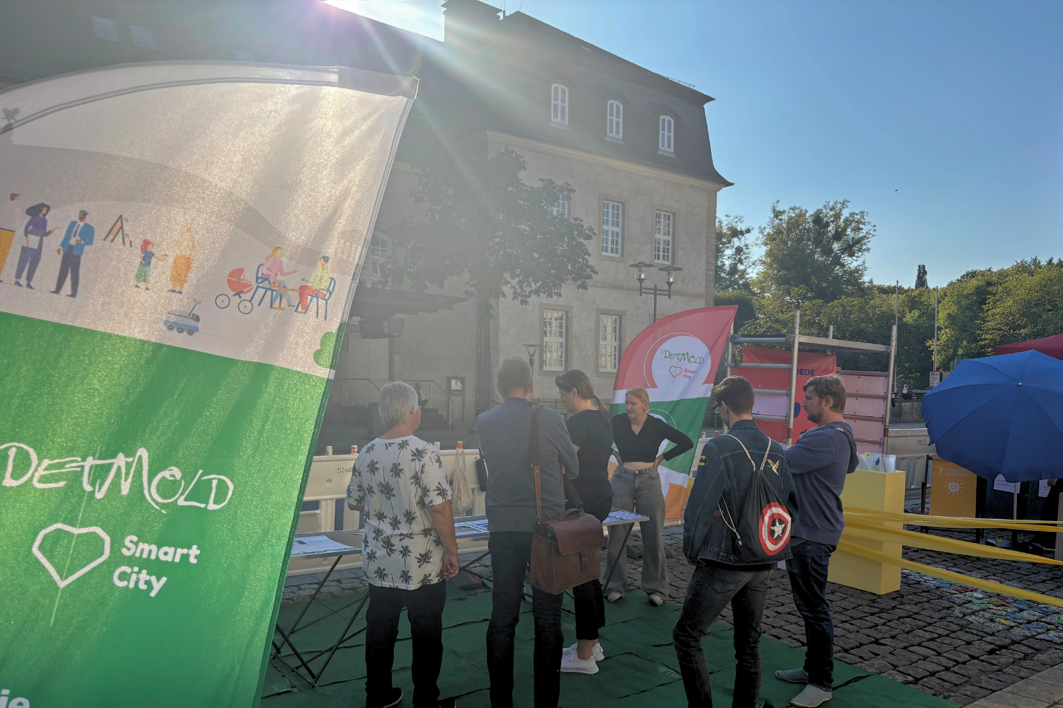 Vorne rechts im Bild sieht man eine grün-weiße Beachflag der Detmold Smart City ins Foto ragen. 5 Besucher*innen informieren sich an einem Tisch zum Projekt bei einer Mitarbeitenden der Stadt Detmold.
