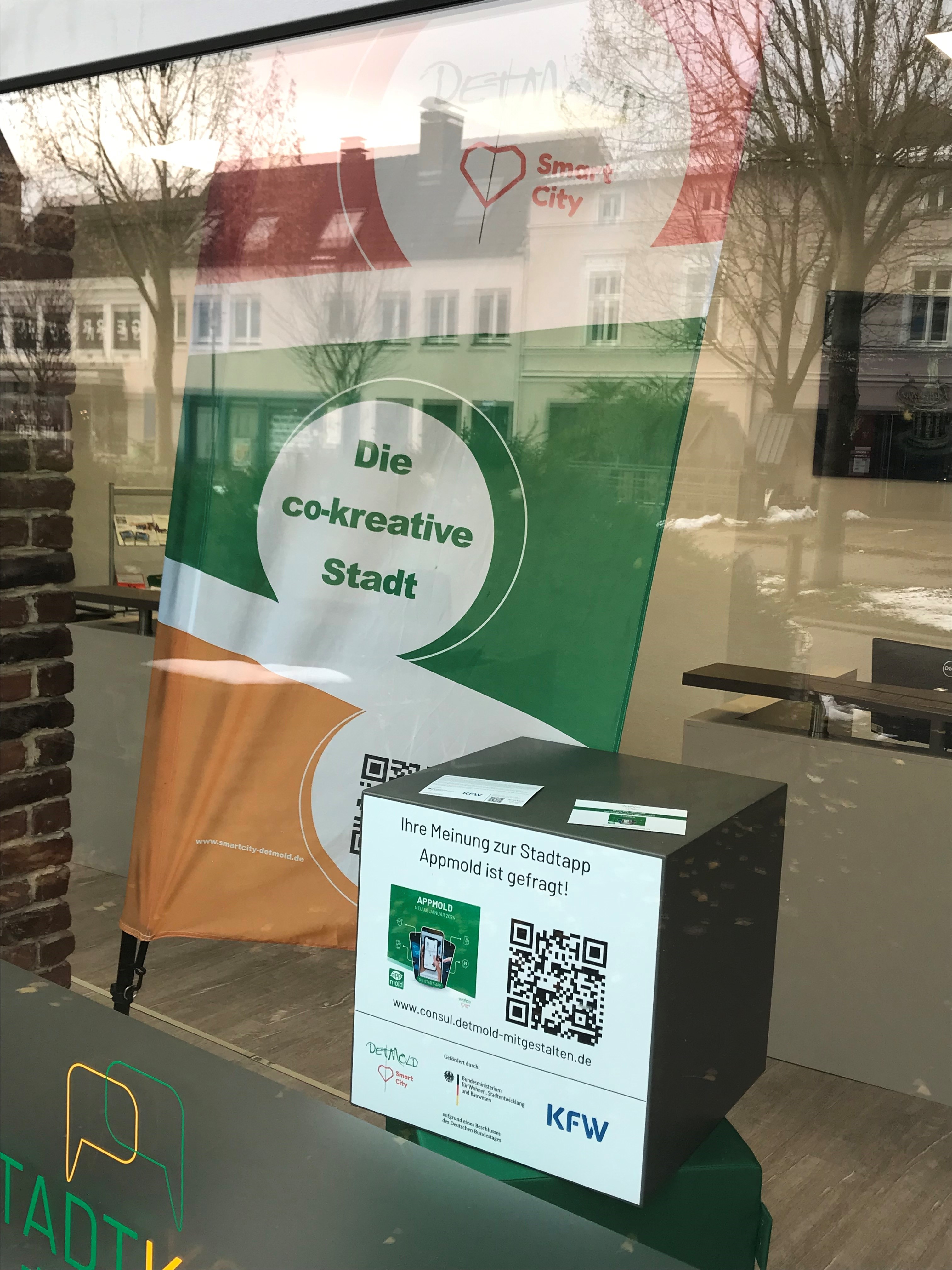 Das Vild zeigt ein Schaufenster in dem eine bunte Beachflag  mit dem Auftrag "Smart City Detmold. Die co-kreative Stadt" zu sehen ist. Davor ist der QR-Code-Würfel platziert, der mit der aufgeklebten QR-Code-Folie auf die Bürgerbeteiligung hinweist.