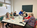 Das Foto zeigt sieben Damen, die um einen weißen Tisch sitzen und sich mit ihren Mobiltelefonen beschäftigen.