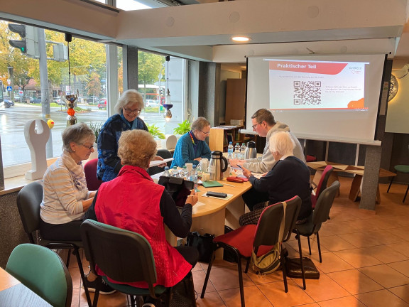 Das Foto zeigt fünf Seniorinnen, die an einem Tisch sitzen und sich autauschen. Oben rechts im Bild ist ein junger Mitarbeiter der Stadt Detmold, der die Seniorinnen schult. Im Hintergrund ist die Präsentationsfläche an der Wand zu sehen sowie eine große Fensterfront. 
