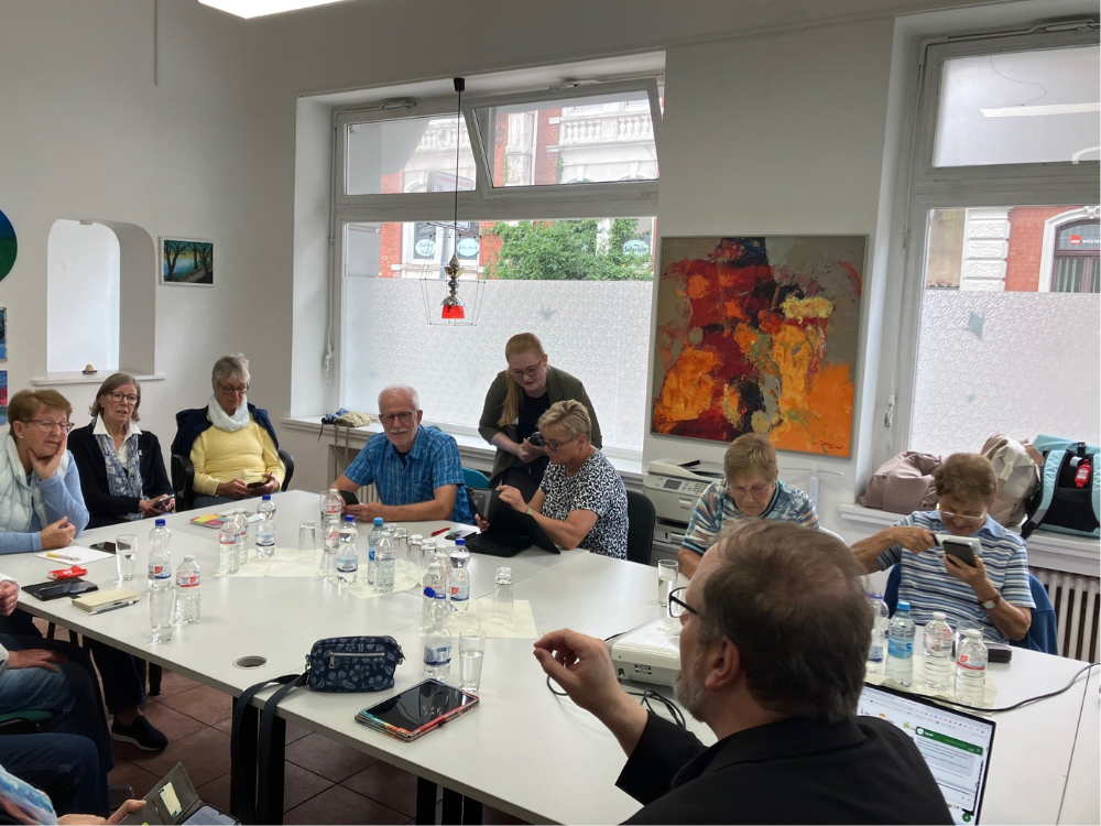 Das Foto zeigt den Seminarraum mit einem großen weißen Tisch und einem großen Fenster im Hintergrund. Die Seniorinnen sitzen und hören dem Vortrag interessiert zu.