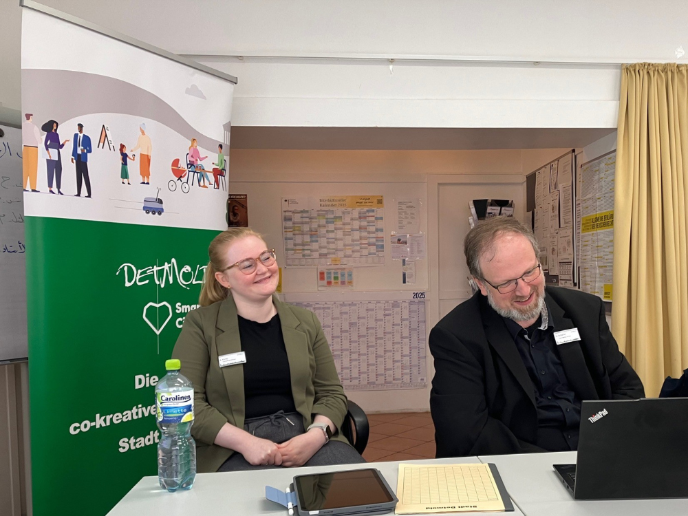 Das Bild zeigt Alina Koray (links) und Dennis Schäffer (rechts) sitzend an einem Tisch, bei der Schulung der Senior*innen. Beide lächeln.