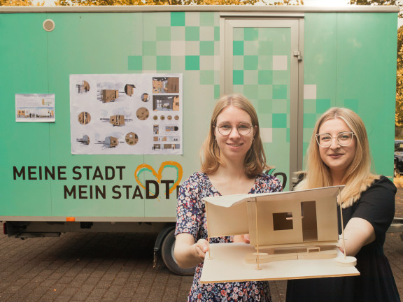 Zwei junge Studentinnen stehen vor einem mintgrünen Anhänger des Detmolder Stadtverkehrs und halten lächelnd ihr Modell eines ausgebauten Anhängers in die Kamera. 