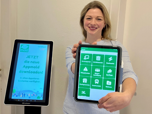 Eine Mitarbeiterin der Stadt steht rechts neben eine Infostele, lächelt und hält ein iPad mit der Stadtapp Appmold in die Kamera. 