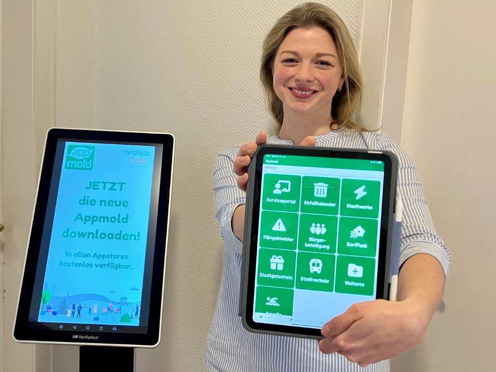 Eine Mitarbeiterin der Stadt steht rechts neben eine Infostele, lächelt und hält ein iPad mit der Stadtapp Appmold in die Kamera.