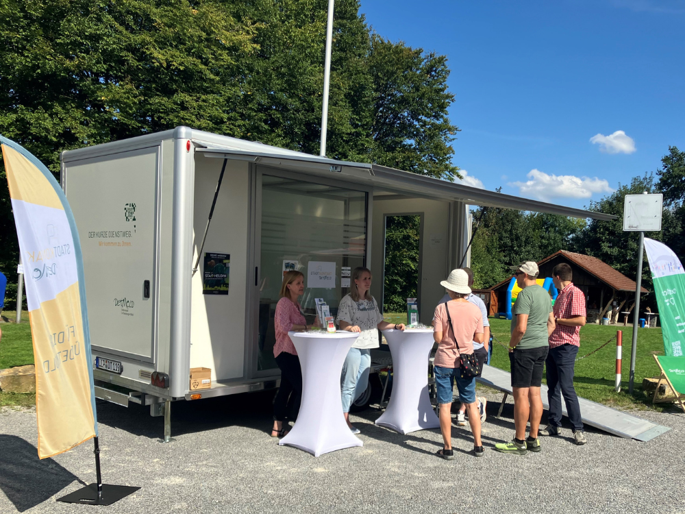 Es ist sehr sonnig. Der städtische Anhänger steht auf einem Schotterplatz, die Klappe ist geöffnet und bietet den beiden städtischen Mitarbeiterinnen Schatten. Sie befinden sich im Gespräch mit einem Ehepaar und einem weiteren Mann.