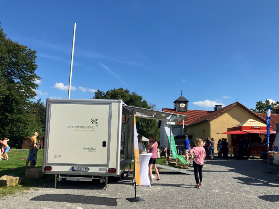 Das Foto zeigt den städtischen Anhänger von hinten, mit ausgeklappter Frontklappe. Es ist ein sionniger Tag und der Anhänger steht auf einem Schotterparkplatz. Mehere Menschen sind (abgewendet von der Kamera) zu sehen. Links im Bild spielen Kinder auf dem Rasen. 