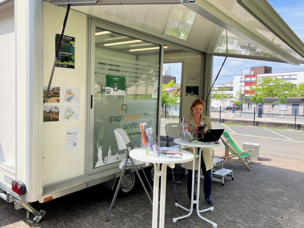 Eine Mitarbeiterin der Stadt Detmold steht vor dem mobilen Anhänger der Stadt an einem Stehtisch, um interessierten Bürgerinnen und Bürgern Rede und Antwort zu stehen.