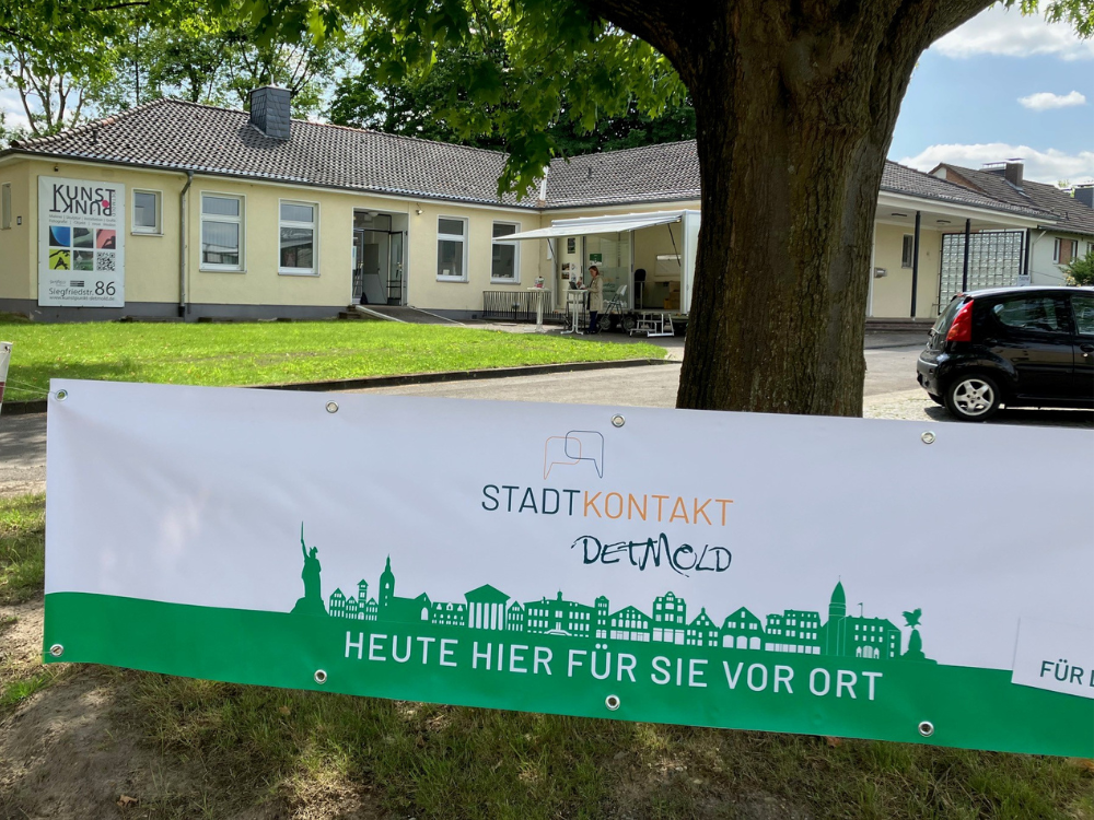 Das Foto zeigt ein Banner, welches zwischen zwei Bäumen aufgehängt ist und auf den Einsatz des Stadtkontakt mobil hinweist. "Heute hier für Sie vor Ort!