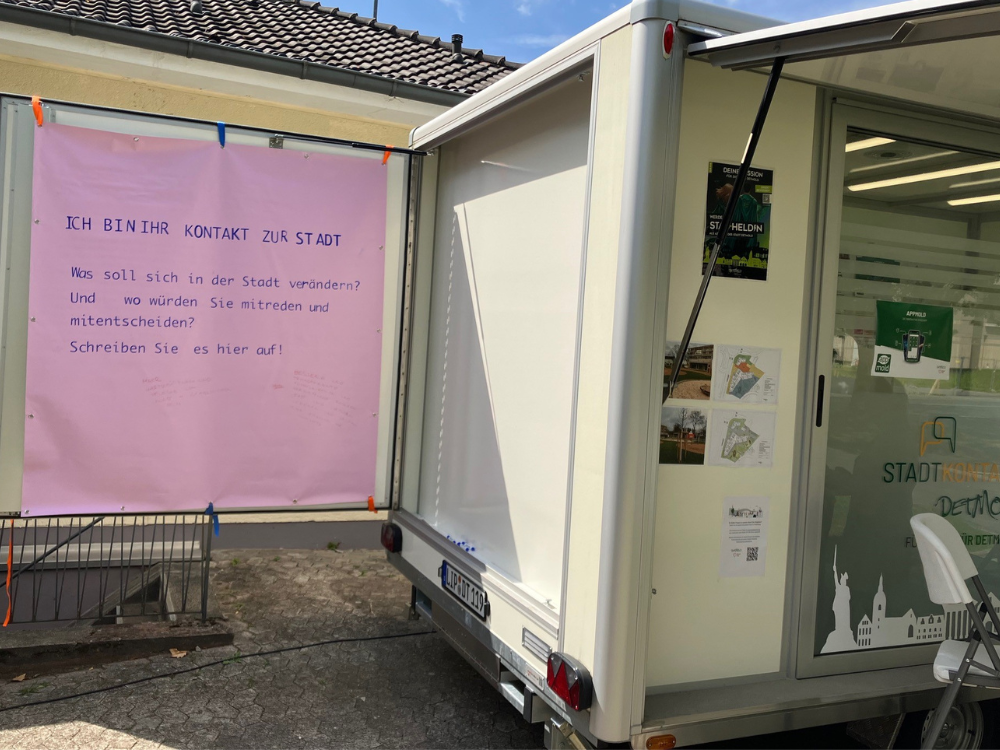 Bürgerbeteiligung mit einem rosa Plakat am Stadtkontakt mobil, das zur Mitgestaltung der Stadt aufruft: 'Ich bin Ihr Kontakt zur Stadt. Was soll sich in der Stadt verändern? Und wo würden Sie mitreden und mitentscheiden? Schreiben Sie es hier auf!'
