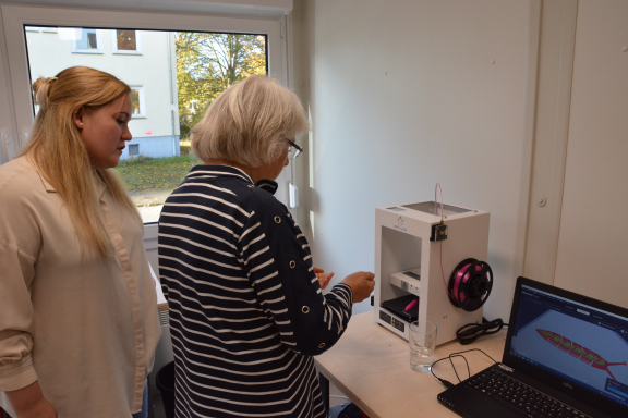 Eine junge Frau steht hinter einer älteren Dame und erklärt ihr die Funktion eines 3D-Druckers, der vor beiden auf einem Tisch steht. 
