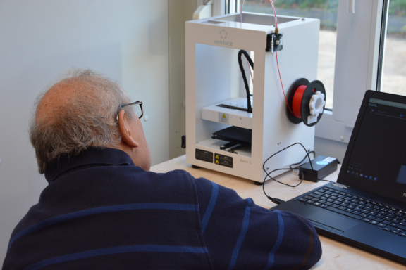 Man sieht einen weißen 3D-Drucker stehend auf einem Tisch. davor sitzt ein älterer Herr auf einem Stuhl und schaut sich interessiert den Druck an. 