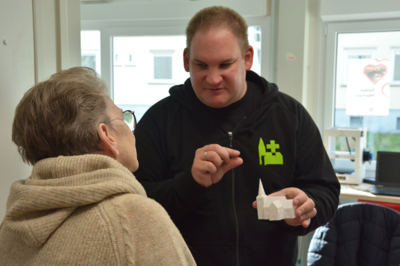 Ein junger Mann im schwarzen Pullover erklärt einer älteren Dame mit Brille ein 3D-Druck-Modell einer Kirche. 