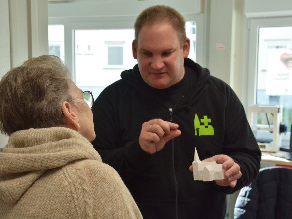 Ein junger Mann im schwarzen Pullover erklärt einer älteren Dame mit Brille ein 3D-Druck-Modell einer Kirche. 