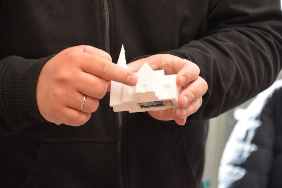 auf dem (angeschnittenen) Foto sieht man die Hände des Dozenten, der ein weißes Modell einer im 3D-Drucker gefertigten Kirche hält. Er trägt einen schwarzen Pullover. 