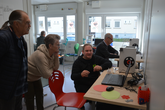 Das Foto wurde in der Mitmachwerkstatt aufgenommen und zeigt eine totale des Raumes, in dem sich 6 Personen an verschiedenen Arbeitstischen befinden. Darauf sieht man 3D-Drucker. 