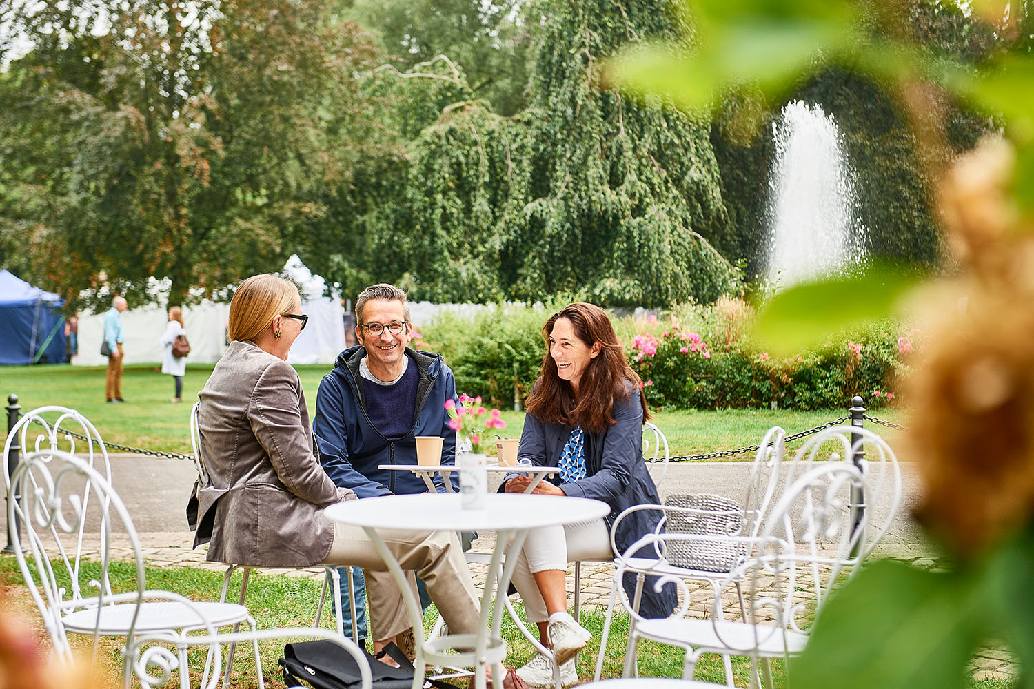 Symbolbild: Zwei Frauen und ein Mann sitzen auf weißen Metallmöbeln in einem Park und unterhalten sihc und lachen. Im Hintergrund ist eine Wasserfontaine zu sehen.