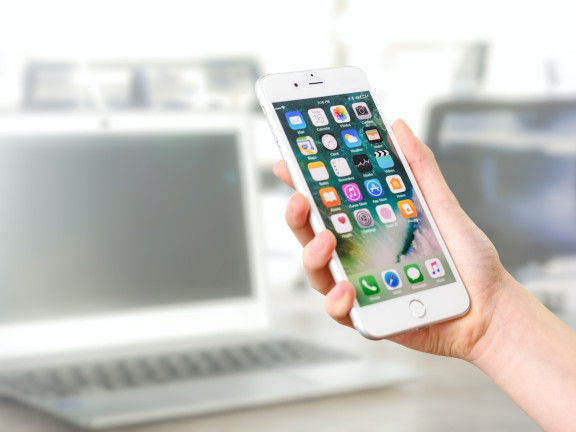 Das Symbolbild zeigt eine Hand, die ein weißes iPhone mit sichtbarem Startbildschirm vor einem unscharfen Laptop im Hintergrund hält. 