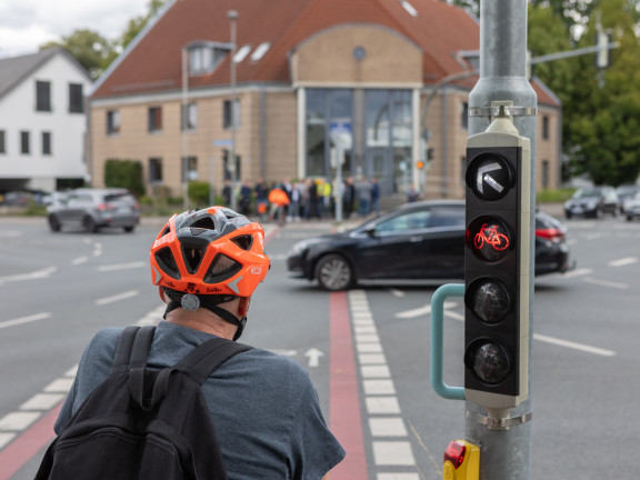 Das Foto zeigt einen älteren Radfahrer von hinten, der an einer belebten Kreuzung an der Ampel steht Er trägt einen rot-schwarzen Helm. Im Hintergrund sind Gebäude, Autos und Menschen zu sehen. 