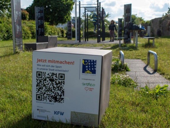 Ein QR-Code-Würfel (grauer Würfel mit 45 cm Kantenlänge und eine Klebefolie, die zur Kamera weist) steht auf einer Rasenfläche. Der QR-Code auf der Folie des Würfels ist gut zu erkennen. 